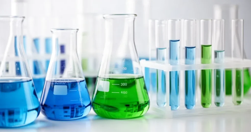 Wzór na stężenie molowe Zbliżenie na szklane naczynia laboratoryjne z kolorowymi roztworami w odcieniach niebieskiego i zielonego na rozmytym tle.